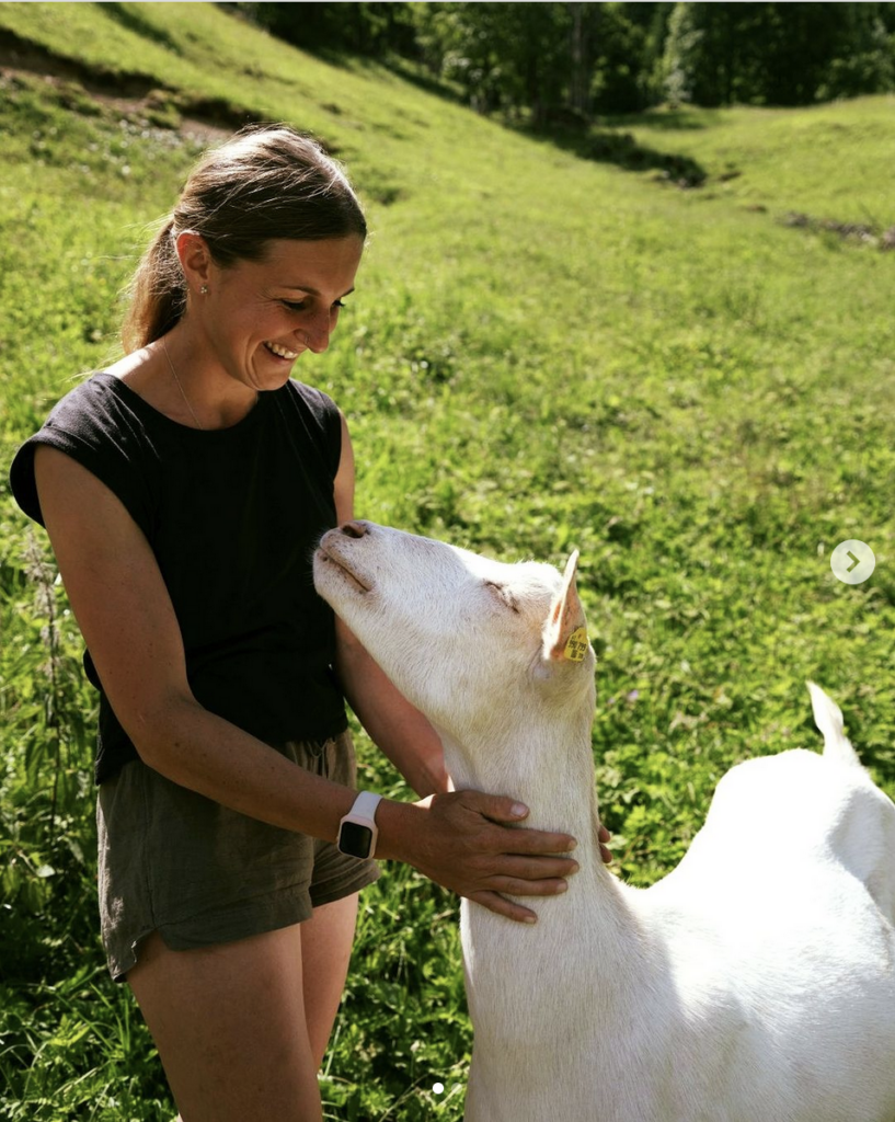 Eine lächelnde Frau auf einer Wiese streichelt den Kopf einer Ziege.